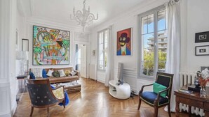Ein geräumiges Wohnzimmer mit Holzboden, einer Couch, einem Sessel und einem Tisch. Große Fenster mit Vorhängen, eine Kristallleuchter und Kunstwerke an den Wänden.