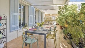Ein Essbereich im Freien mit Tisch und Stühlen, umgeben von Topfpflanzen und einer überdachten Terrasse.
