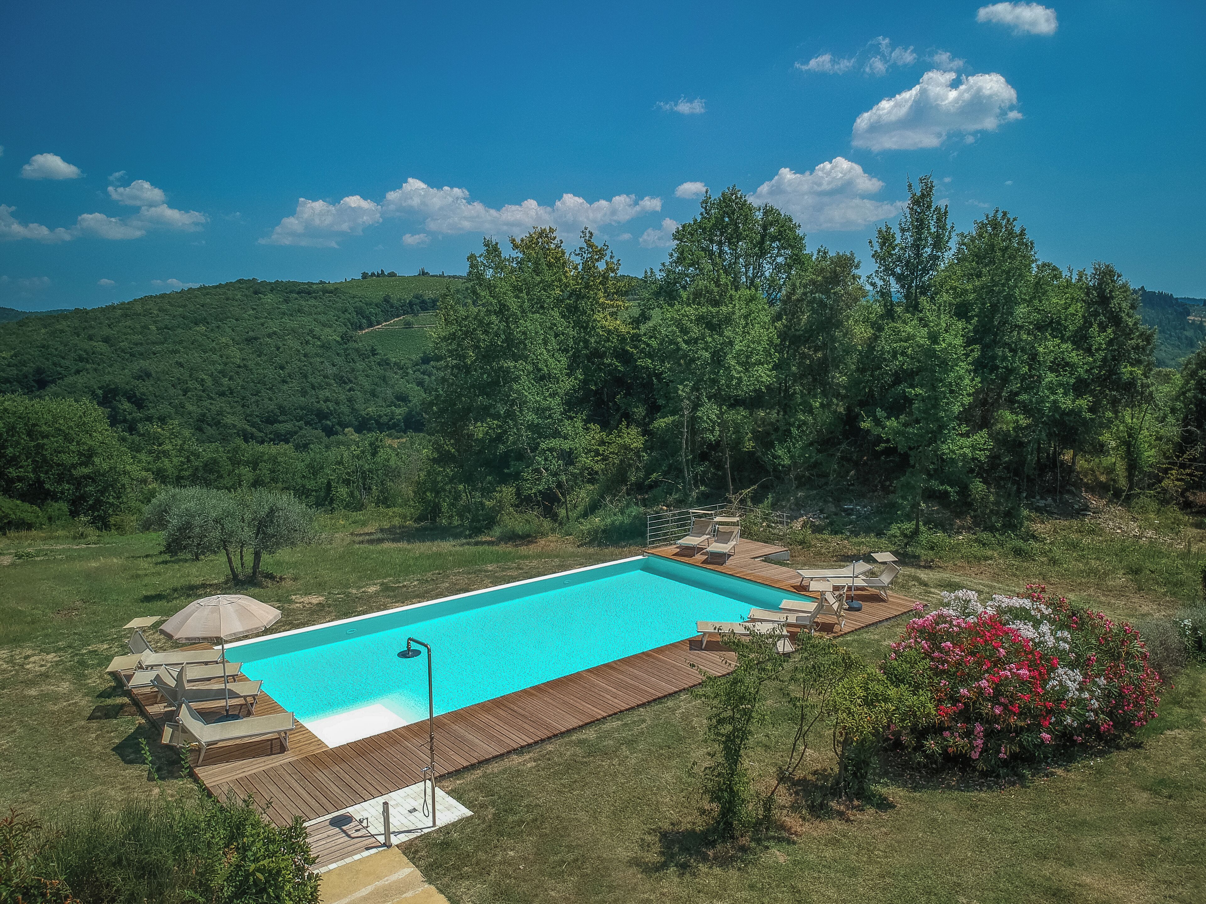 A rectangular swimming pool with a wooden deck and lounge chairs, surrounded by lush greenery and trees.