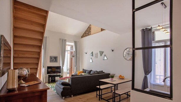 A modern living room with a wooden staircase, a sofa, a dining table, and a large window with curtains. - Villa Sandrine (Calvi)