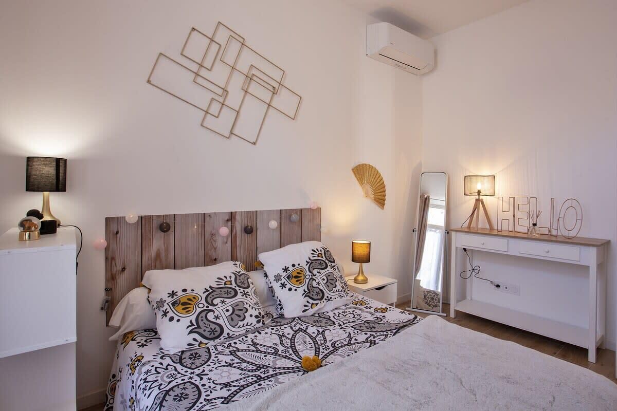 A bedroom with a wooden headboard, patterned bedding, a bedside table with a lamp, a mirror, and a dresser with a 'HELLO' sign.