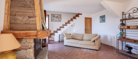 A living room with a stone fireplace, a wooden staircase, a beige sofa, and a tiled floor.