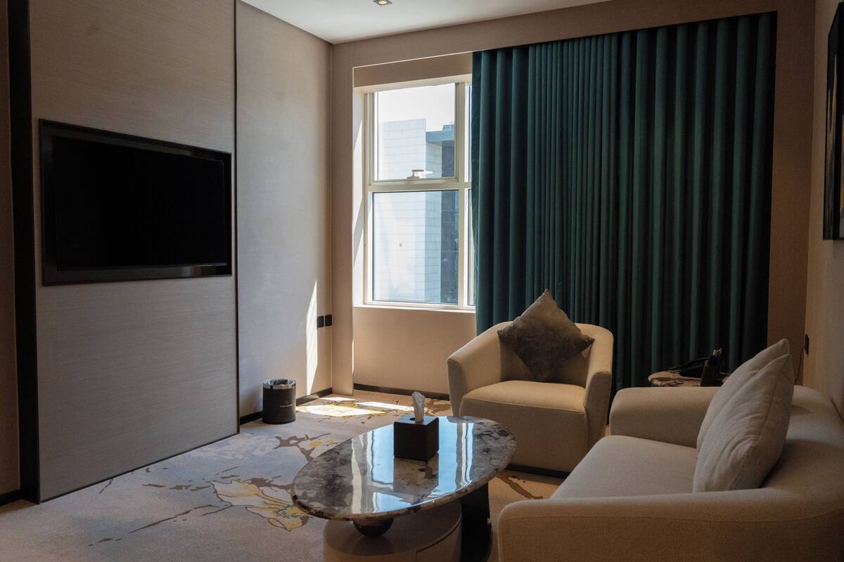 A modern hotel room with a flat-screen TV mounted on the wall, a sofa, a coffee table, and a large window with curtains.