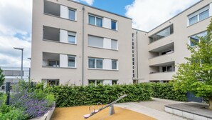Un immeuble moderne à plusieurs étages, doté de balcons, avec une balançoire dans la cour et un ciel dégagé.