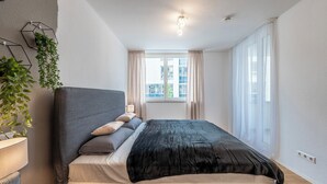 Une chambre à coucher moderne avec un grand lit, une table de chevet, une lampe et une fenêtre avec des rideaux.