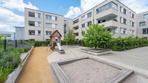 Un terrain de jeux avec un toboggan et un bac à sable, entouré d’un jardin aménagé et d’un immeuble à plusieurs étages doté de balcons.