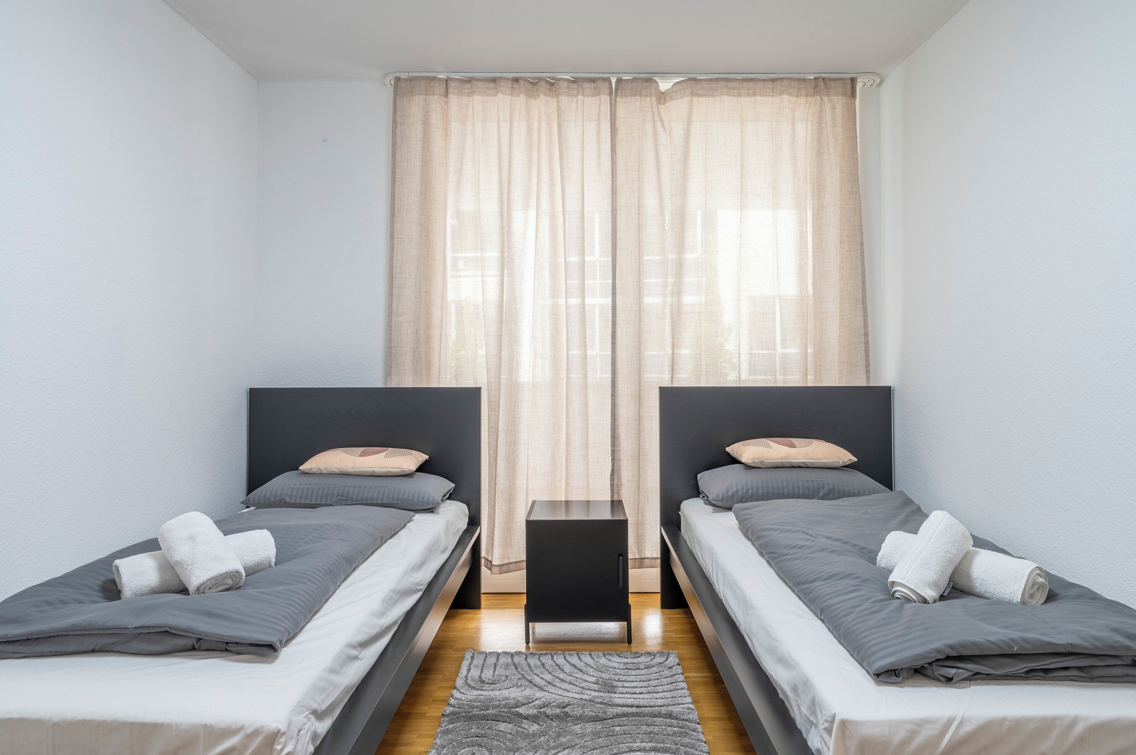 A twin room with two single beds, a small black nightstand, and a large window with sheer curtains.