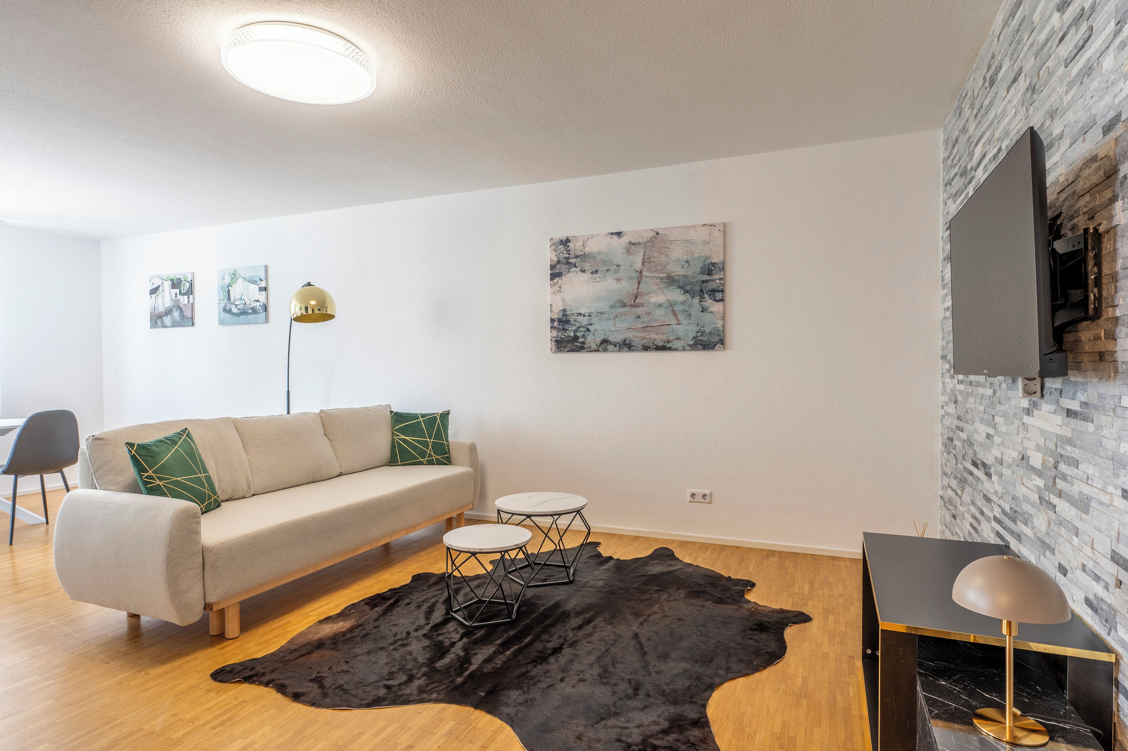 A modern living room with a sofa, two side tables, a black leather rug, a flat-screen TV mounted on a brick wall, and artwork on the wall.