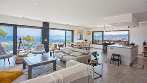 A modern living room with a sofa, chairs, and a dining area. There is a large window offering a view of the sea and mountains.