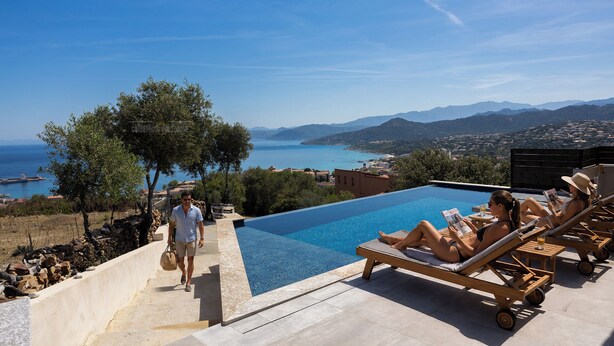 A poolside area with lounge chairs, a pool, and a view of the sea and mountains.