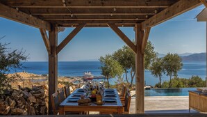 A wooden pergola with a table set for dining, overlooking a coastal view with a ship docked at a pier.