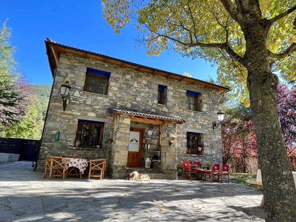 A stone building with a wooden porch, a table with chairs, and a dog in front. - Casa Rural Hostel dos Lucas (OROS BAJO)