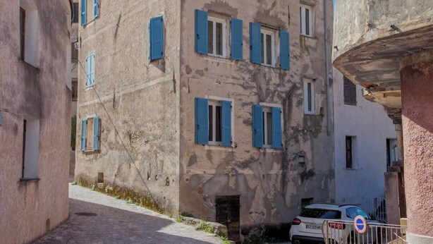 A narrow street with a building featuring blue shutters and a white car parked in front. - Casa Rione (Omessa)