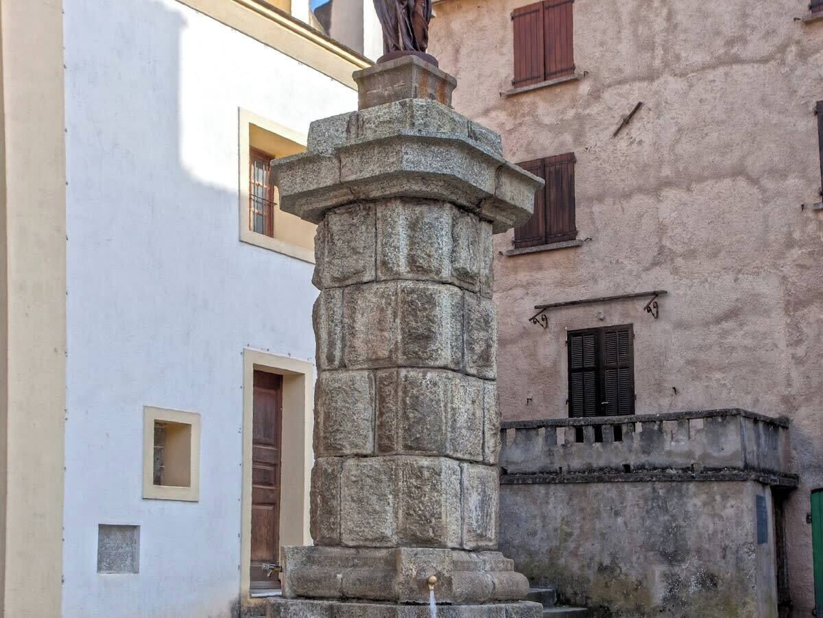 A stone monument with a statue on top, surrounded by buildings with windows and doors.