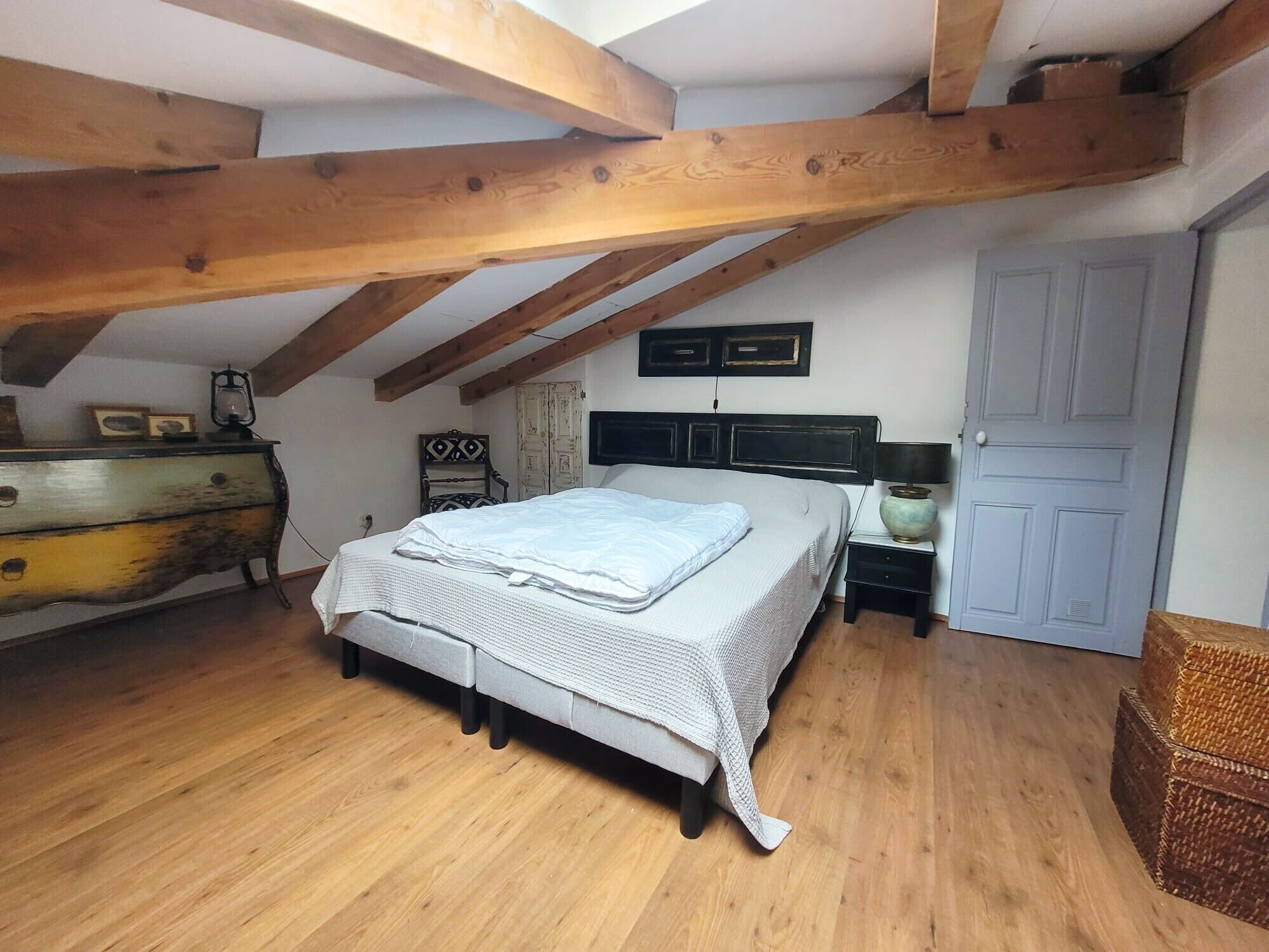 Ein Schlafzimmer mit schrägen Wänden, Holzbalken, einem Bett, einem Schminktisch, einem Sessel, einer Lampe und einer Tür.