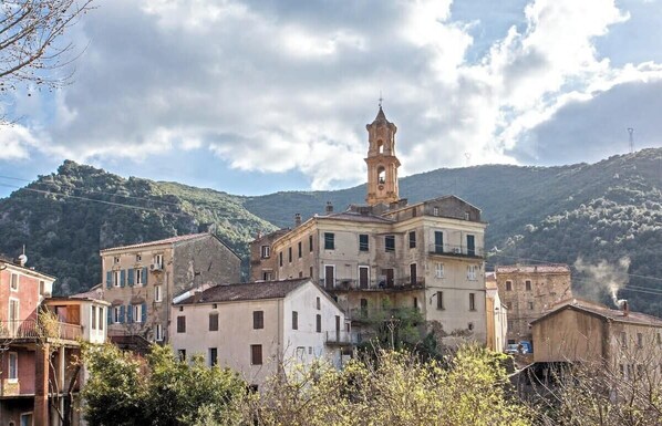 A historic town with a prominent bell tower, surrounded by multi-story buildings with balconies and a mountainous backdrop. - Casa Torra. (Omessa)