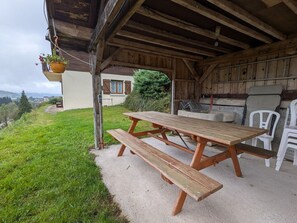 Struttura in legno rustica con tavolo e sedie da picnic, una pianta pensile e vista su una casa sullo sfondo.