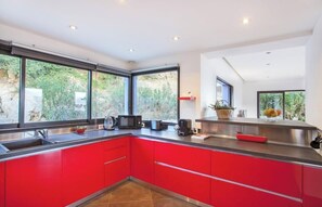 A modern kitchen with red cabinets, a stainless steel countertop, and large windows offering a view of greenery. - Villa Annonciade – Vue panoramique sur l'Île-Rousse, piscine chauffée (Ile-Rousse)