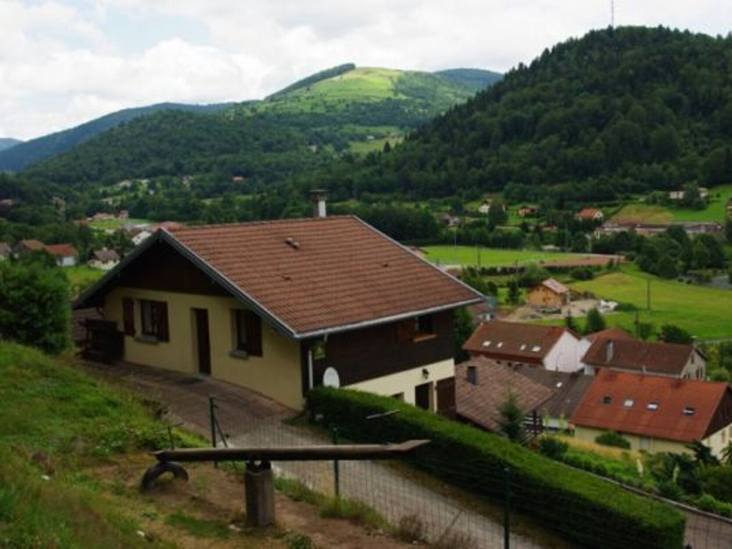 A rural house with a sloped roof, surrounded by lush greenery and hills in the background.