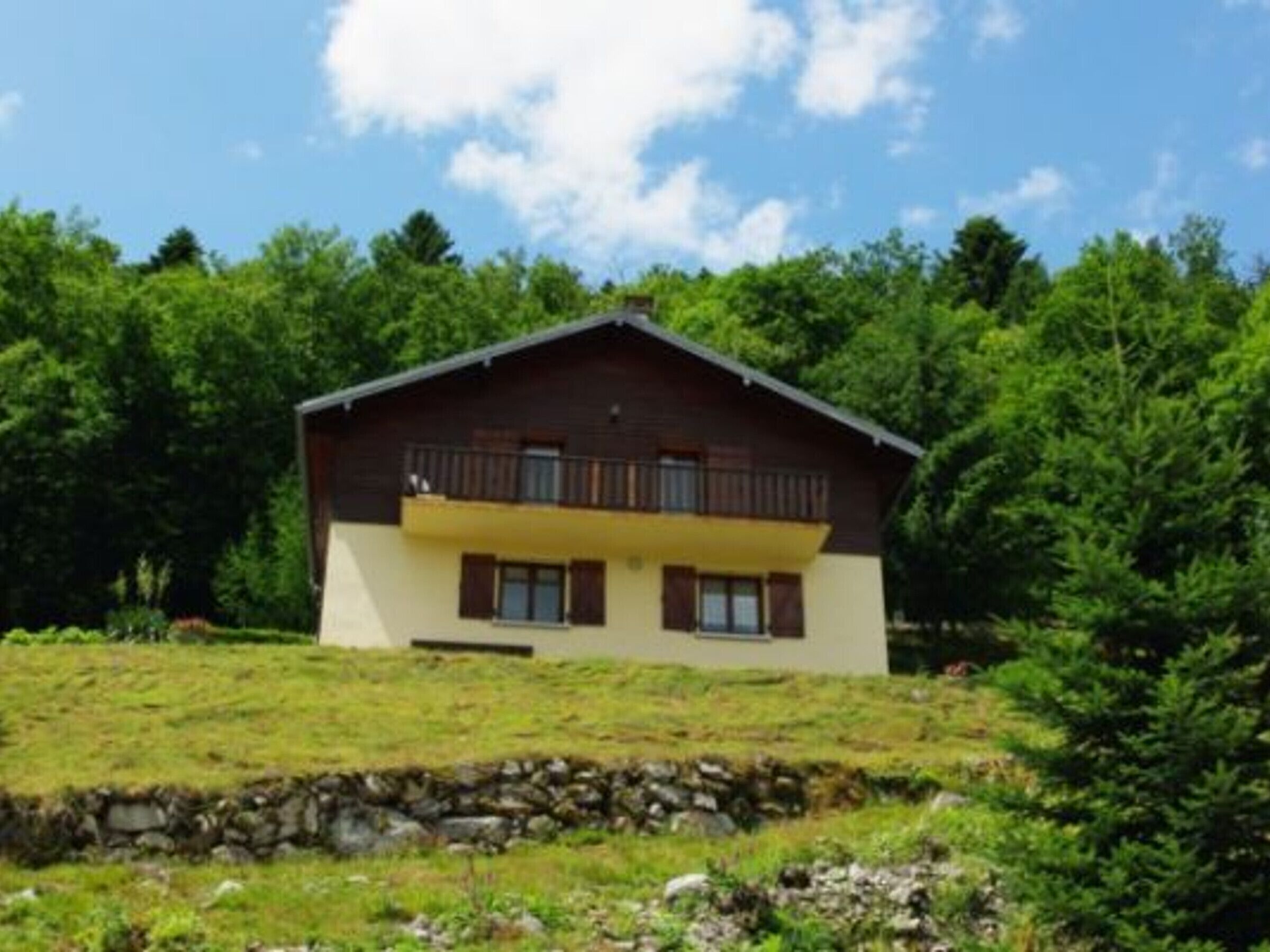 A two-story chalet with a balcony, surrounded by lush greenery and a stone wall.