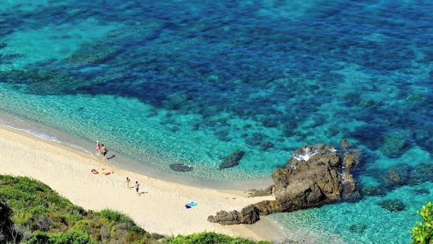 Una playa con agua turquesa cristalina, afloramientos rocosos y gente caminando por la orilla.