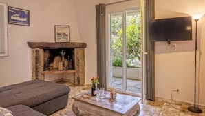 Una sala de estar con chimenea de piedra, un sofá gris, una mesa de centro con vasos y una botella, un televisor de pantalla plana y un ventanal con cortinas.