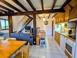 Uma cozinha acolhedora com vigas de madeira, uma área de refeições com mesa e cadeiras, uma escada que leva a um loft, e um balcão com eletrodomésticos.