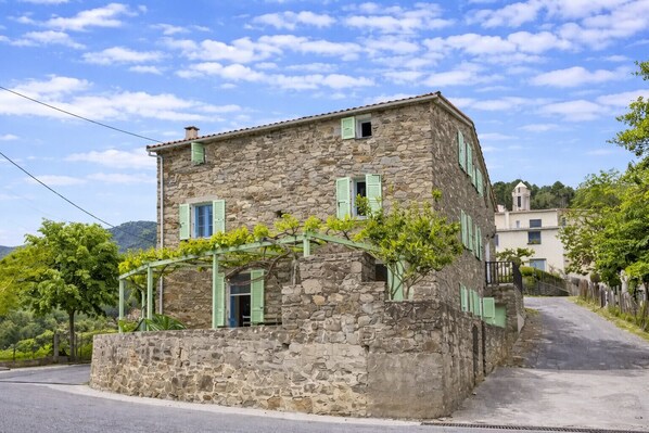 A stone building with a sloped roof, green shutters, and a vine-covered pergola.