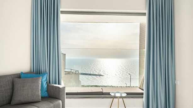 A modern living room with a grey sofa, a small round table, and a view of a body of water through a large window. - Netanya Beach & Pool Stay by Sea N' Rent (Netanya)