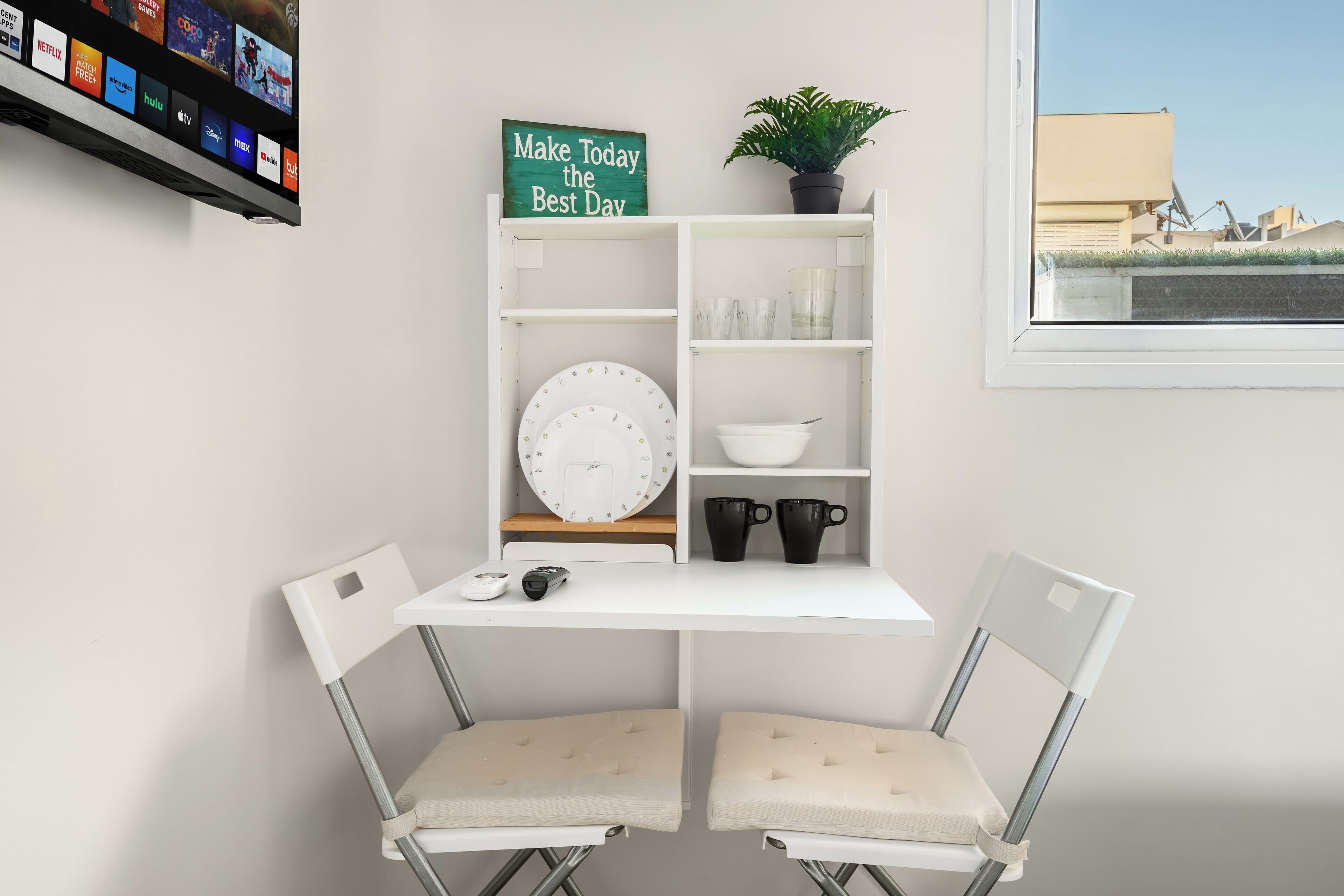 Quarto moderno com escrivaninha branca, duas cadeiras, um relógio, um vaso de planta e uma janela com vista para a cidade.