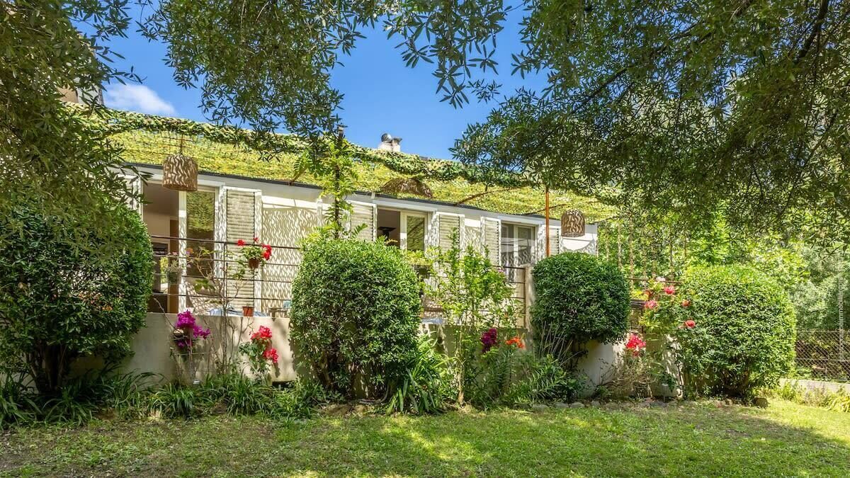 A house with a green roof, surrounded by lush vegetation and flowering plants.