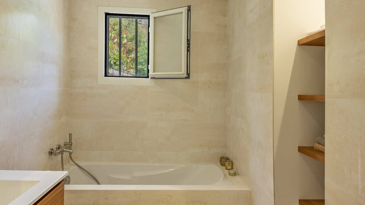 A bathroom with a bathtub, a window with a view of greenery, and wooden shelves.