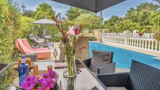 Zona junto a la piscina con mobiliario de mimbre, una silla colgante y una mesa con bebidas y flores.