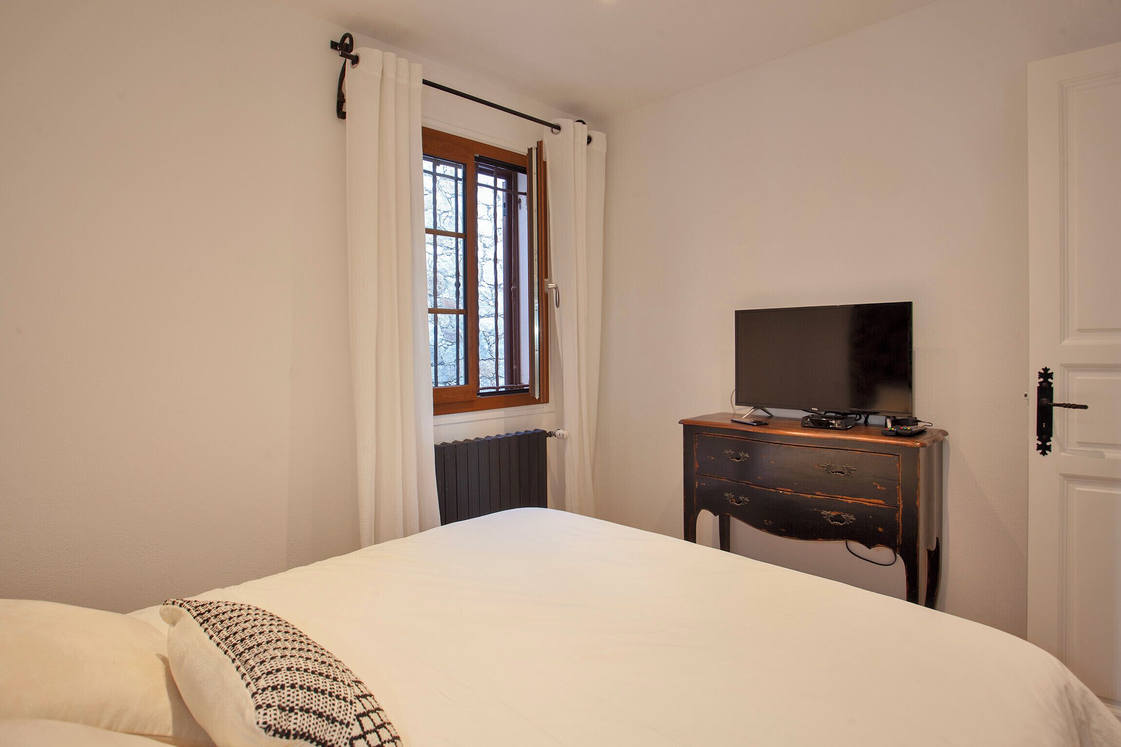 Een slaapkamer met een bed, een kaptafel met een televisie, een raam met gordijnen en een radiator.