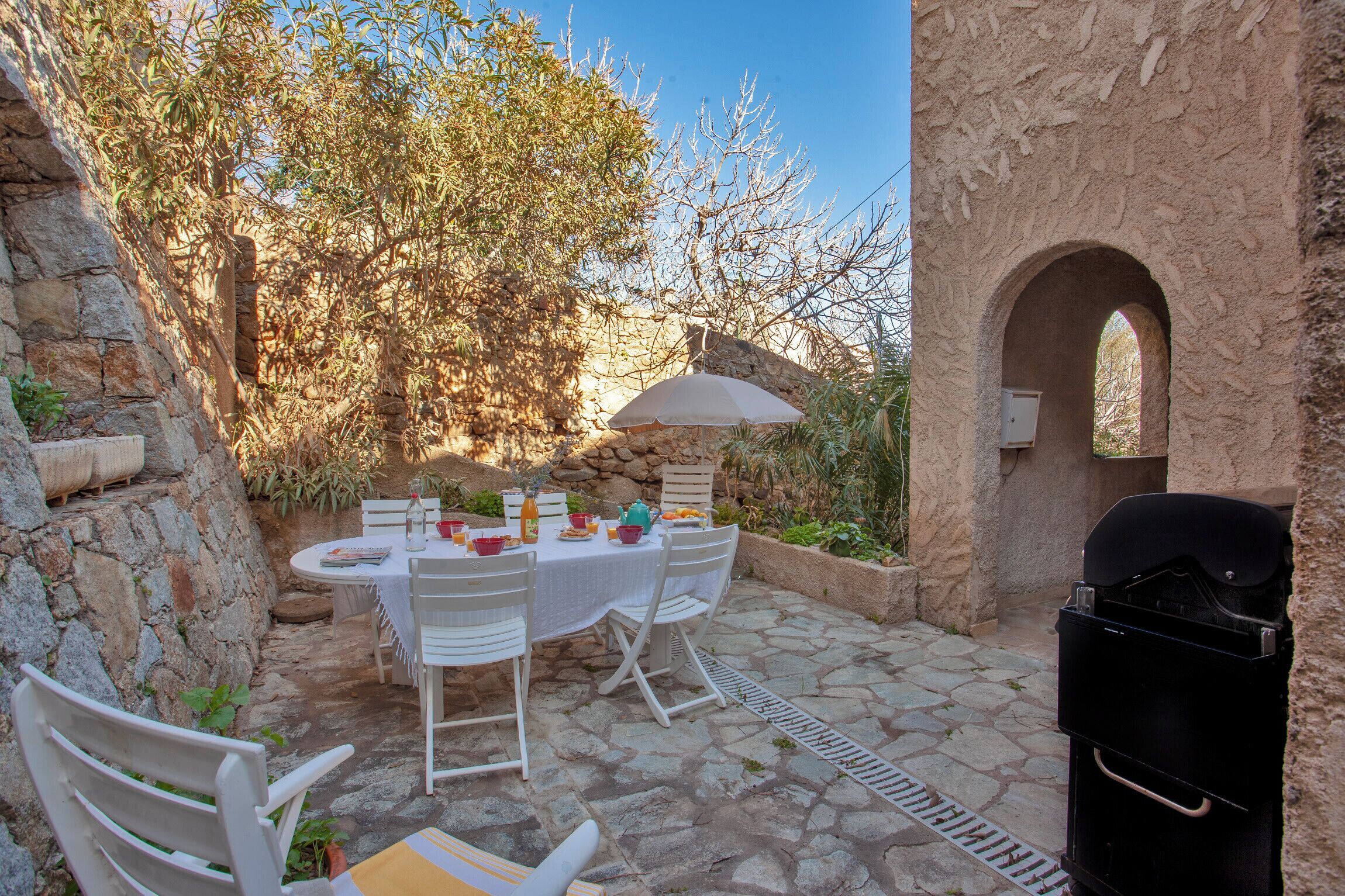 Un espace extérieur aménagé pour les repas, avec des murs en pierre, une table dressée pour deux personnes et un barbecue.