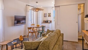 A modern living room with a sofa, dining table, and chairs. There is a TV mounted on the wall and a kitchen area in the background. - Casa Félicie (Calvi)