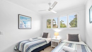 A bedroom with two beds, a ceiling fan, a window with blinds, and a framed picture on the wall.