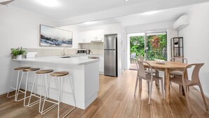 A modern kitchen with white cabinetry, a central island with stools, a dining area with a wooden table and chairs, and a view of greenery outside.