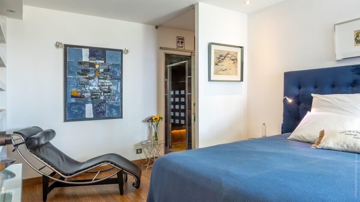 A modern hotel room with a bed, a massage chair, a small table with a vase, and artwork on the walls.