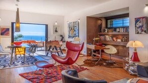 A modern living room with a view of the ocean, a dining area with a table and chairs, and a bookshelf filled with books. - Casa Di A Scala (Brando)