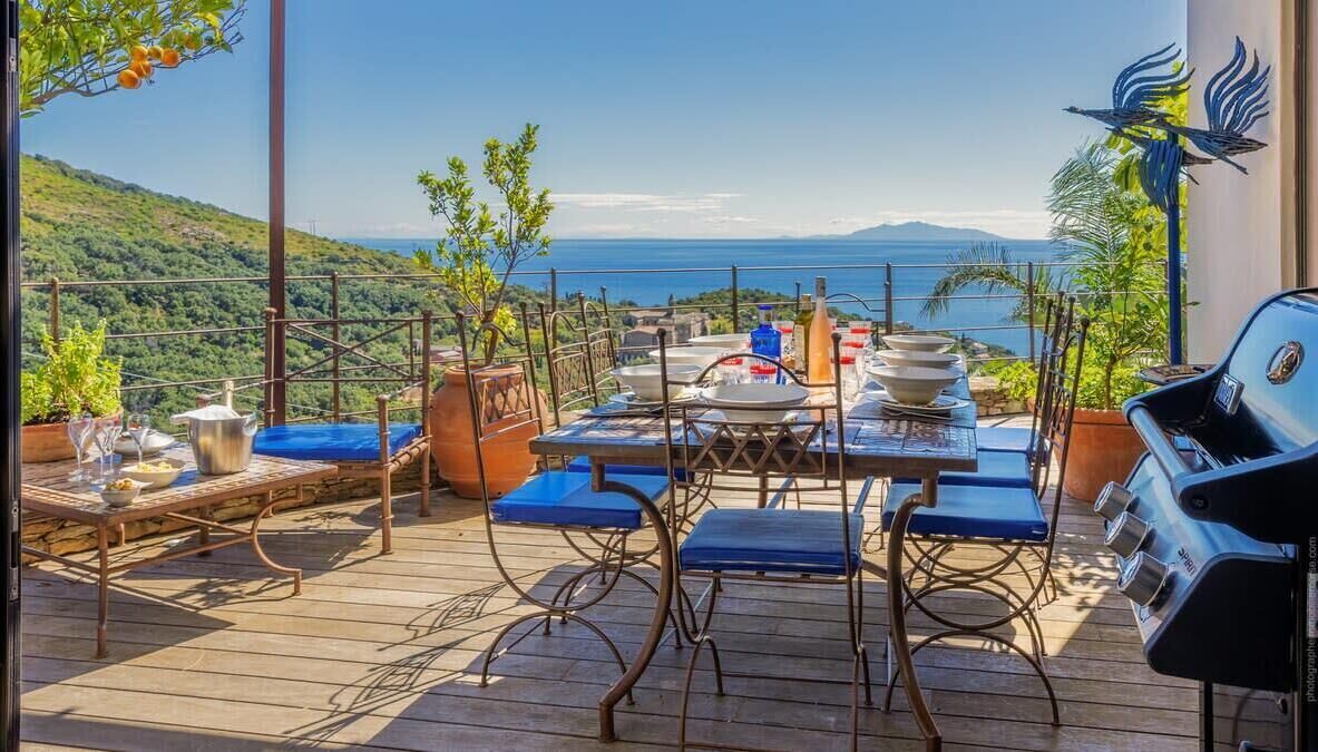 A wooden deck with a table and chairs, a grill, and a view of the sea and hills.