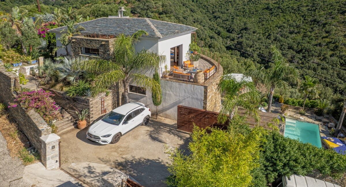 Une maison moderne, avec une voiture blanche garée devant, entourée d’une végétation luxuriante et dotée d’une piscine.