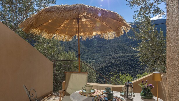 A terrace with a thatched umbrella, a table with a cup, a vase with flowers, and a view of a forested valley.