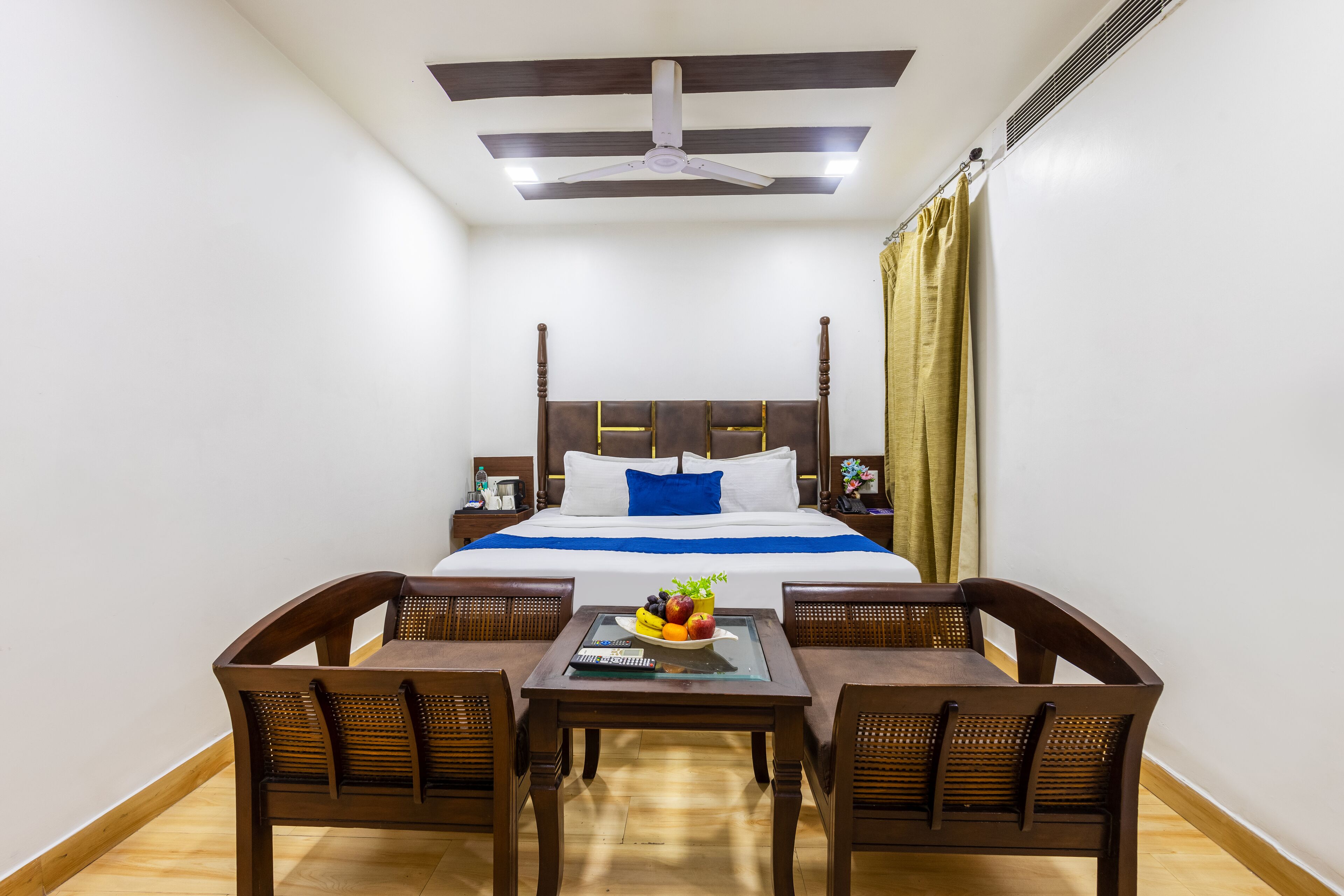 Une chambre d’hôtel avec un lit, une table avec des fruits, deux chaises, un ventilateur de plafond et une table de chevet avec une lampe.