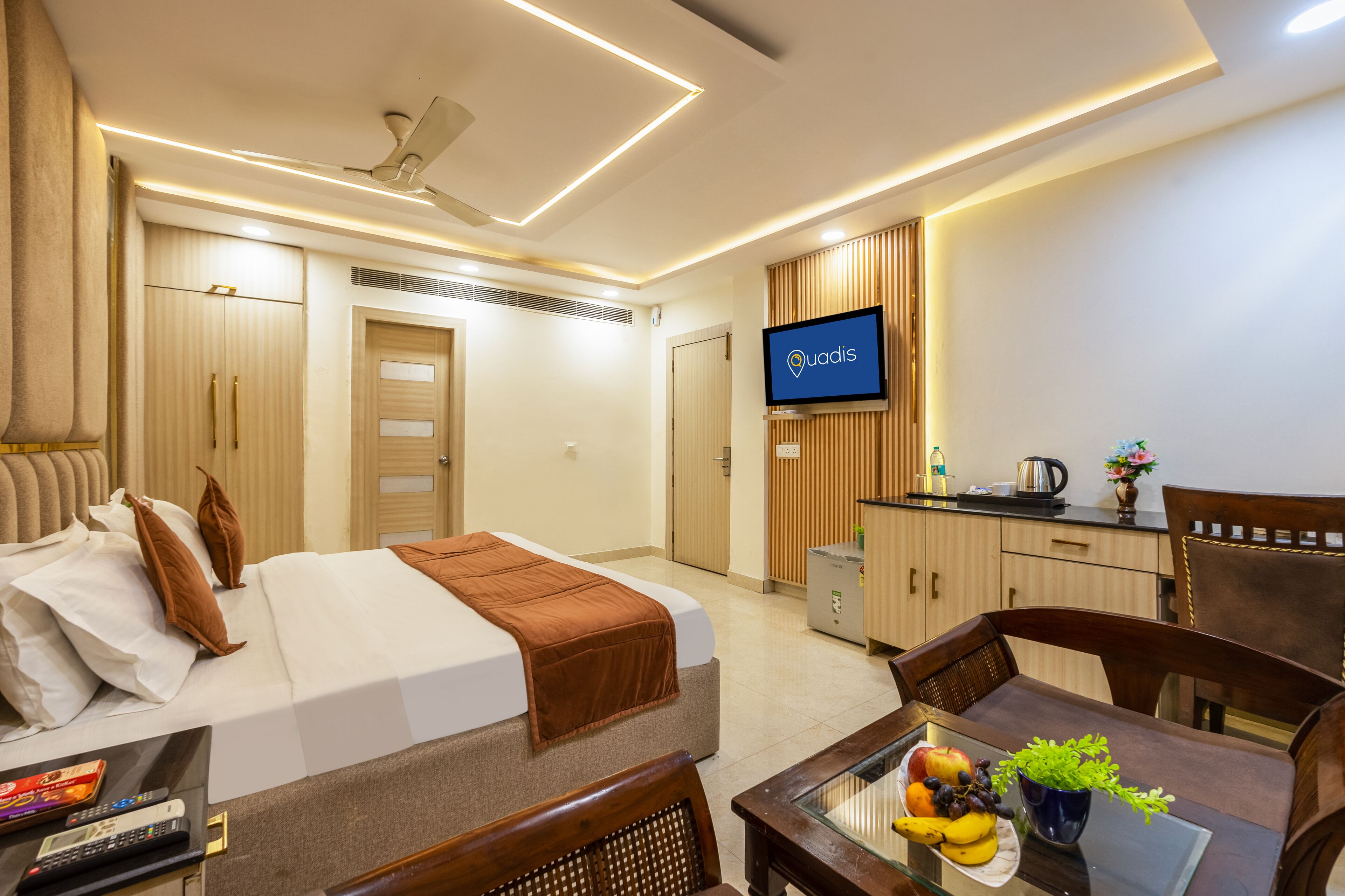 A hotel room with a bed, a TV, a dining table with fruit, and a kitchenette.