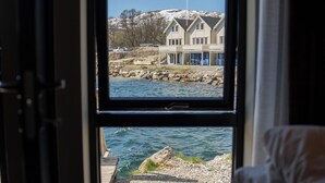 A room with a bed, a window with a view of a house and water. - Steam Pier Adventure Hotel (Tromsø)