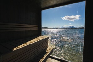A sauna with a wooden bench, a large window, and a view of a lake with mountains in the distance. - Steam Pier Adventure Hotel (Tromsø)