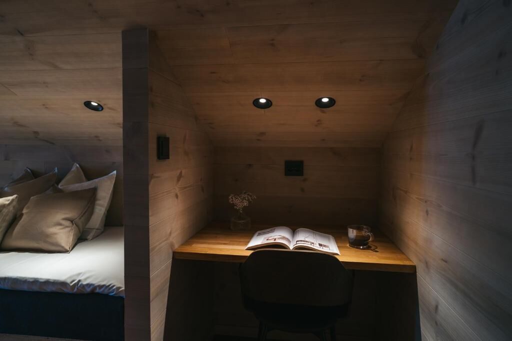 Ein kleiner, gemütlicher Raum mit einem Bett, einem Schreibtisch und einem Stuhl. Auf dem Schreibtisch liegen ein Buch und ein Glas.