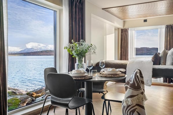 A room with a dining table set for two, a sofa, and a view of a lake and mountains. - Steam Pier Adventure Hotel (Tromsø)