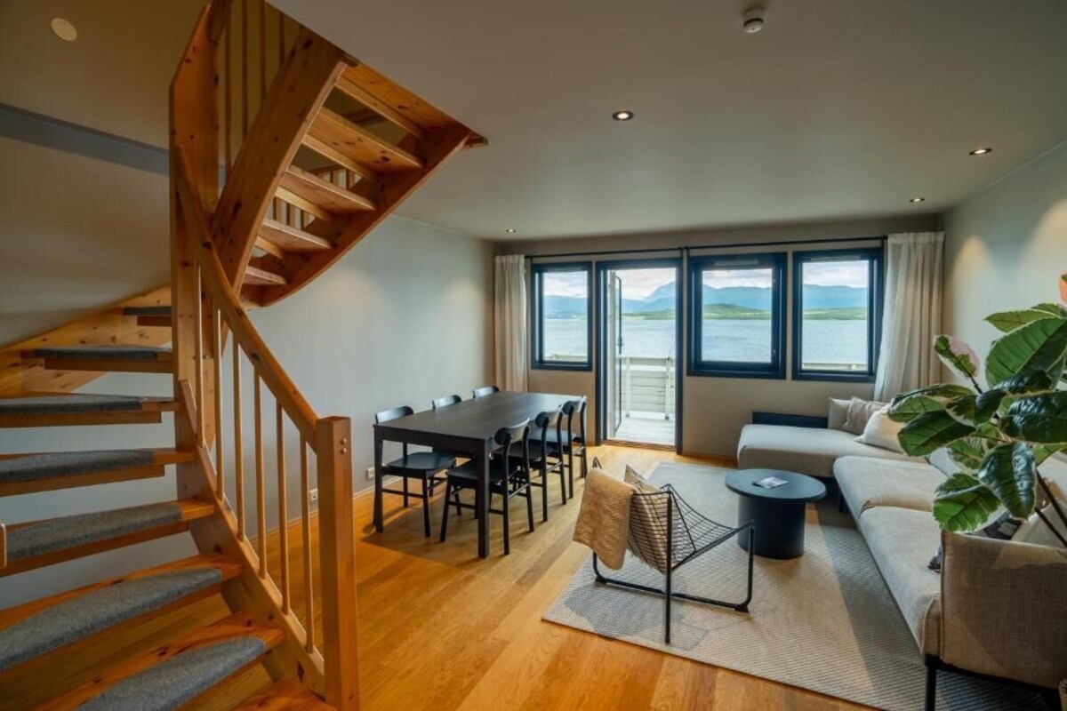 Ein geräumiges Wohnzimmer mit einer Holztreppe, einem Esstisch mit Stühlen, einer Couch und einem großen Fenster mit Blick auf das Wasser.